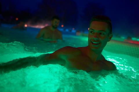 Junger Mann im Außenbecken am Abend bei Dunkelheit, das Wasser wird grün angestrahlt