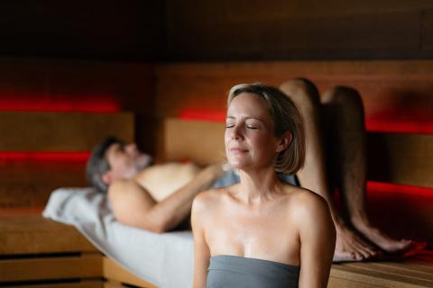 Eine Frau sitzt in der Sauna. Sie hat die Augen geschlossen und genießt den Moment. Hinter ihr liegt ein Mann in der Sauna.